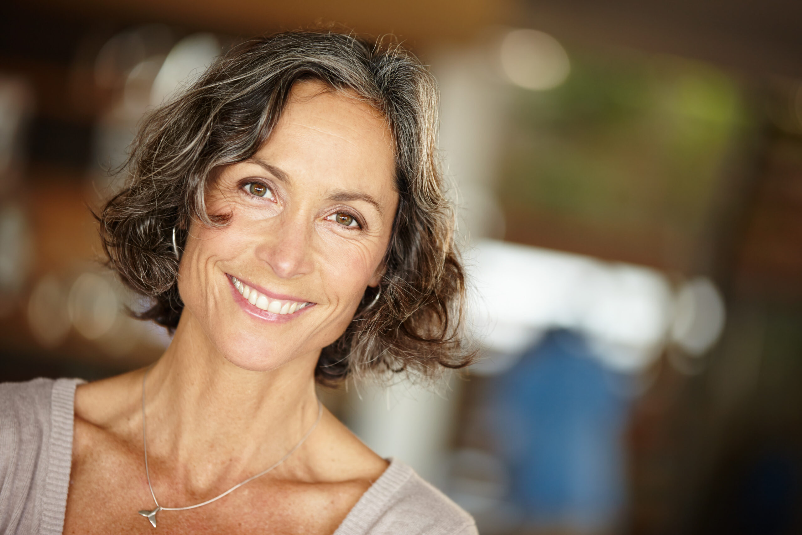 A middle-aged woman with wavy brown and gray hair smiles at a med spa in Hayden, Idaho, wearing a light sweater and necklace. at North Idaho Med Spa in Hayden