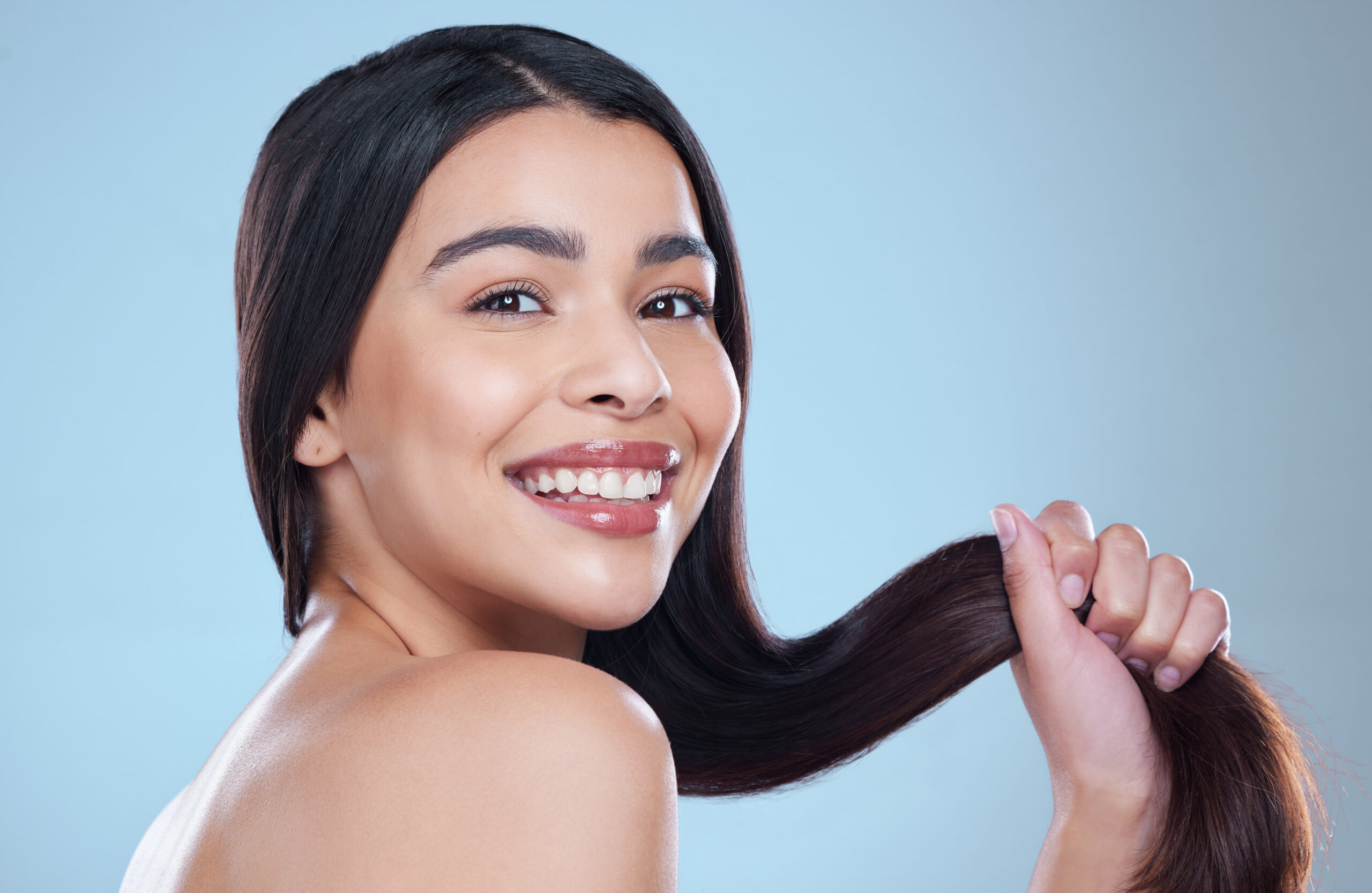 A confident, smiling woman with long dark hair holds her hair against a light blue background, highlighting Hayden Idaho med spa results. at North Idaho Med Spa in Hayden